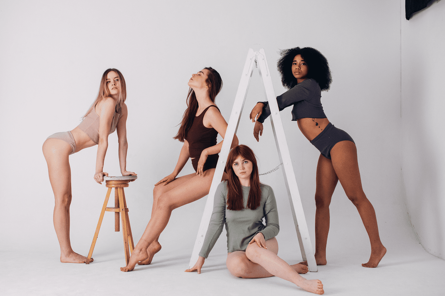 Four women posing in a studio, using a stool and ladder as props, in minimalistic attire.